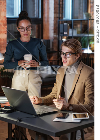 Handsome young man in glasses using laptop while presenting project to manager with coffee cup in dark office 89070033