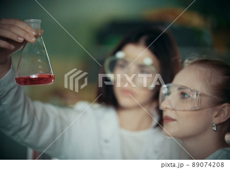 Chemical laboratory. Two young woman holding a flask with red liquid in it Chemical laboratory. Two young woman holding a flask with red liquid in it 89074208