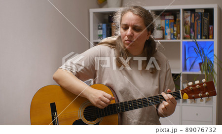 A girl with a serious expression on her face plays an acoustic guitar while sitting in a room. Behind her is a rack of board games and flowers. Musical composition. 89076732