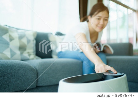 Happy Asian young woman turning on high efficiency air purifier while staying and reading a book in the living room. Woman using air purifier in her house. 89076747