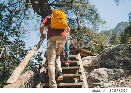 Rear view young adult man climbing a wooden ladder in the mountains. Casually dressed tourist with yellow backpack going up the wooden crafted ladder in the national park with big boulders and cliffs 89078292