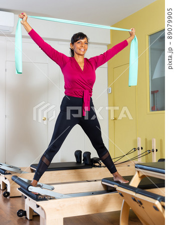 Positive Hispanic woman performing stretching pilates exercises on reformer with elastic band Positive Hispanic woman performing stretching pilates exercises on reformer with elastic band 89079905