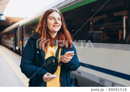 Young redhhead woman waiting on station platform with backpack on background electric train using smart phone. Railroad transport concept, Traveler Young redhhead woman waiting on station platform with backpack on background electric train using smart phone. Railroad transport concept, Traveler 89081518