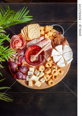 Round cutting board with slicing assorted cheese, sausage rosette, ham, cracker, grapes and figs with honey. Charcuterie and cheese platter. Top view 89082234