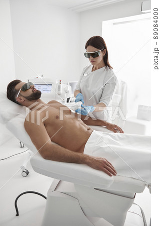 Young master with goggles conducts laser epilation to bearded man client lying on couch in clinic 89084000