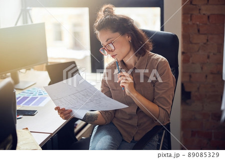 Focused young woman designer holding and studying blueprint paper while working on new interior design project in her office 89085329