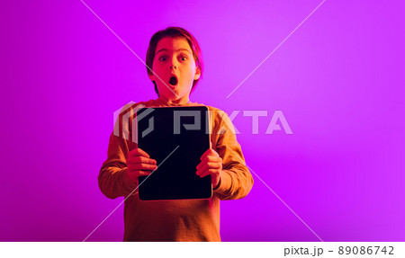 Portrait of boy, child with expressive emtions holding tablet isolated over pink background in neon light 89086742