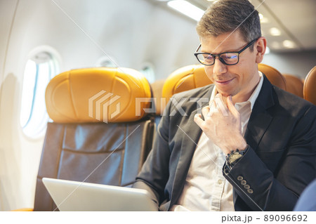 During a flight, a businessman in formal attire and glasses looks out the plane window while working on a laptop computer. Service with Internet access on board and a business travel concept 89096692