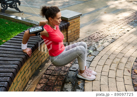 Sportive active middle-aged African American woman exercising outdoor. Active sportswoman doing triceps exercises leaning her hands on a wooden bench in a city park 89097725
