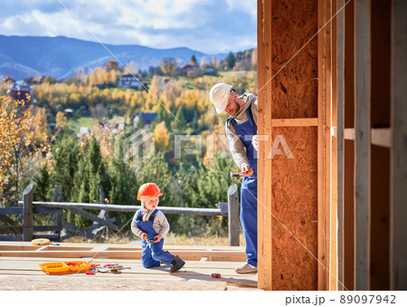 Father with toddler son building wooden frame house. Male builders hammering nail into plank on construction site, wearing helmet and blue overalls on sunny day. Carpentry and family concept. 89097942