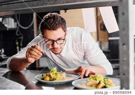 Young serious chef in eyeglasses decorating main dish on plate on counter before delivery Young serious chef in eyeglasses decorating main dish on plate on counter before delivery 89098094