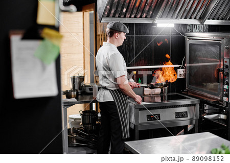 Young chef in apron using his skills to cook dish on open fire in kitchen of restaurant Young chef in apron using his skills to cook dish on open fire in kitchen of restaurant 89098152