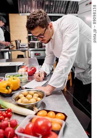 Young chef making a menu at table with food ingredients during his work in kitchen of restaurant Young chef making a menu at table with food ingredients during his work in kitchen of restaurant 89098158