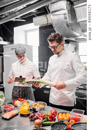 Young chef in uniform standing in kitchen and checking quality of fresh vegetables on table before cooking Young chef in uniform standing in kitchen and checking quality of fresh vegetables on table before cooking 89098173