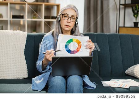Attractive pleasant mature female sitting on sofa and showing graphs and charts. 89099558