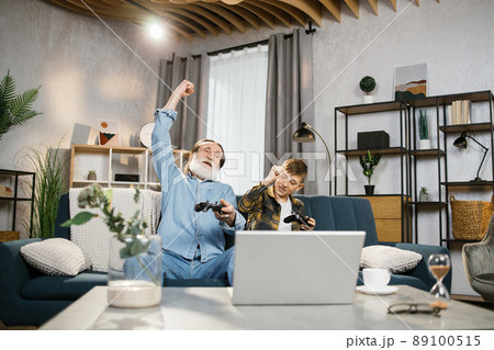 Elder man and his grandson sitting on couch and using wireless joysticks for playing video games Elder man and his grandson sitting on couch and using wireless joysticks for playing video games 89100515