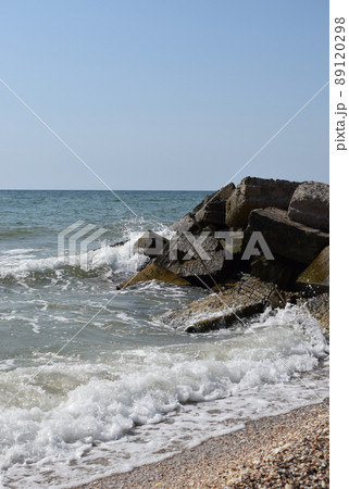 The waves of the turquoise sea break on the coastal boulders and a dense white foam is formed. Clear blue sky. White waves breaking on sandy beach through gap in rocks on sea 89120298