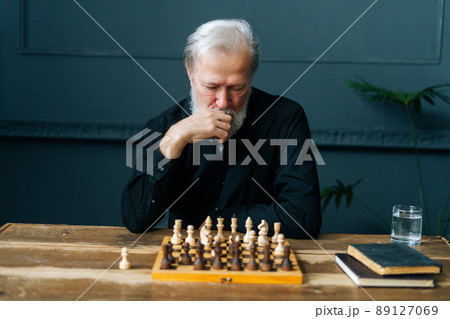 Front view of thoughtful bearded mature pensioner male thinking about chess move sitting at home and playing chess alone. Pensive senior adult man thinking game strategy at table with chessboard. 89127069