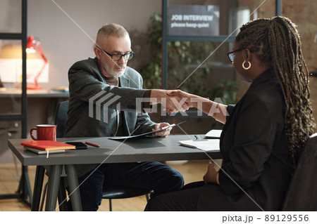 Mature manager shaking hands with young woman while they sitting at table during job interview Mature manager shaking hands with young woman while they sitting at table during job interview 89129556