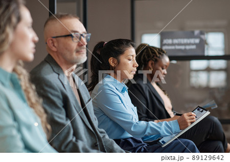 Young serious businesswoman filling resume form for job interview while sitting in waiting room with people Young serious businesswoman filling resume form for job interview while sitting in waiting room with people 89129642