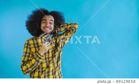 A young man with an African hairstyle on a blue background looks at the phone and is happily surprised. Emotions on a colored background A young man with an African hairstyle on a blue background looks at the phone and is happily surprised. Emotions on a colored background 89129658