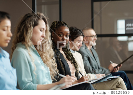 Portrait of African young woman looking at camera while filling application among candidates for job interview Portrait of African young woman looking at camera while filling application among candidates for job interview 89129659