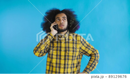 A young man with an African hairstyle on a blue background is talking on the phone. On a colored background A young man with an African hairstyle on a blue background is talking on the phone. On a colored background 89129676