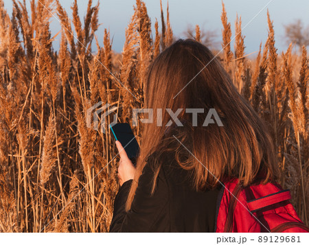 Pretty young woman in black coat with backpack smartphone on pampas grass sunset sky dry reeds Millennial long hair girl Pretty young woman in black coat with backpack smartphone on pampas grass sunset sky dry reeds Millennial long hair girl 89129681