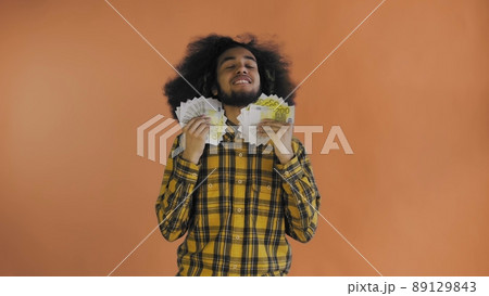A happy African-American man holding a fan of banknotes in his hands and looking at the camera, standing isolated on an orange background 89129843
