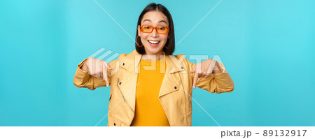 Portrait of stylish asian girl, wears sunglasses, smiles and points fingers down, shows advertisement, stands over blue background Portrait of stylish asian girl, wears sunglasses, smiles and points fingers down, shows advertisement, stands over blue background 89132917