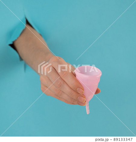 A female hand sticking out of a hole from a blue background holds a pink menstrual cup.  89133347