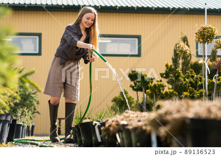 Happy female garden center worker watering plant pots with a hose Happy female garden center worker watering plant pots with a hose 89136523