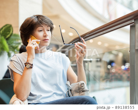 Woman is talking by smartphone and is looking surprisingly on her eyeglasses at shopping mall. Coffee break at store. 89138555