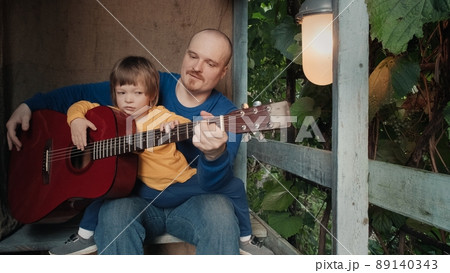 Father with a small child sits on the porch of an old house and plays an acoustic guitar for his cute baby. The concept of family pastime, relationships and teaching children music Father with a small child sits on the porch of an old house and plays an acoustic guitar for his cute baby. The concept of family pastime, relationships and teaching children music 89140343