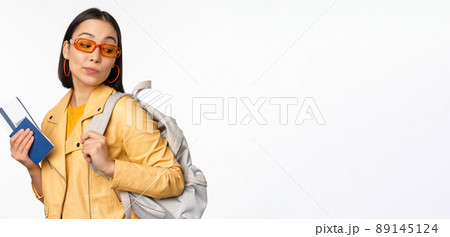 Stylish asian woman in sunglasses going abroad with backpack, tourist holding passport and flight boarding tickets, standing over white background 89145124