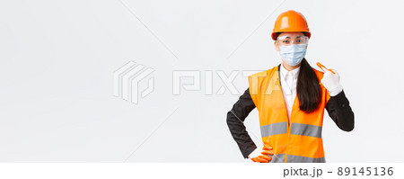 Covid-19 safety protocol at enterpise, construction and preventing virus concept. Confident asian female engineer in helmet and glasses pointing finger at face mask during coronavirus pandemic 89145136