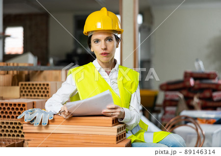 Woman foreman in a yellow vest and a protective helmet keeps track of bricks 89146314