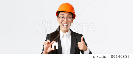 Close-up of upbeat professional female asian engineer, architect in helmet and business suit encourage contact her company for renovation and design, showing house and thumbs-up 89146352