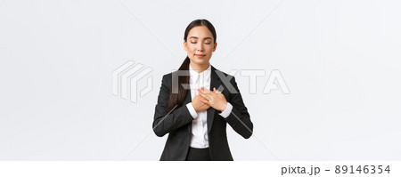 Happy dreamy asian female entrepreneur in suit close eyes and holding hands on heart, remember lovely memory, thinking about something pleasant over white background 89146354