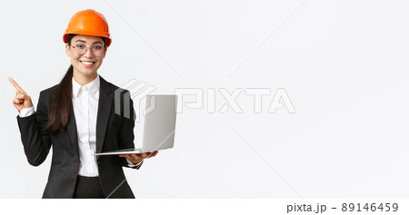 Smiling professional asian female engineer or architect at construction, wearing safety helmet and suit, pointing finger left while using laptop computer, standing white background Smiling professional asian female engineer or architect at construction, wearing safety helmet and suit, pointing finger left while using laptop computer, standing white background 89146459