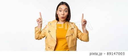 Surprised asian girl pointing fingers up, express interest, showing advertisement ahead, demonstrating promo banner on top, standing against white background Surprised asian girl pointing fingers up, express interest, showing advertisement ahead, demonstrating promo banner on top, standing against white background 89147003