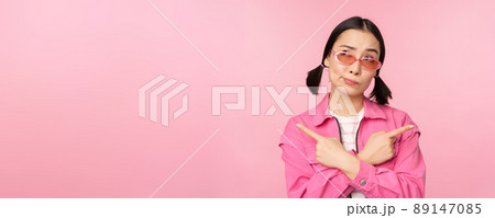 Choice. Stylish korean girl, asian female model points fingers sideways, shows two variants, product advertisement, demonstrating items, standing over pink background Choice. Stylish korean girl, asian female model points fingers sideways, shows two variants, product advertisement, demonstrating items, standing over pink background 89147085