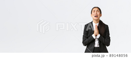 Overworked and distressed asian businesswoman begging for help, screaming hold hands together, pleading and crying desperate, standing upset over white background 89148056