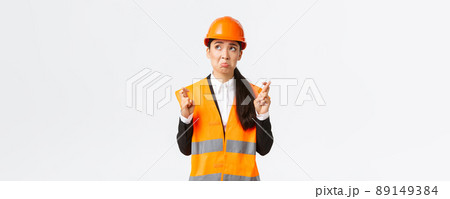 Sad nervous and unconfident asian female construction manager in safety helmet, pouting and look in panic upper left corner, making wish, cross fingers good luck, pleading over white background 89149384