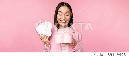 Happy cute korean girl in suit, opens up heart shaped box with romantic gift on white day holiday, standing in suit over pink background 89149408