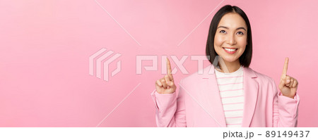 Smiling korean businesswoman, pointing fingers up, showing advertisement, banner or logo on top, standing in suit over pink background Smiling korean businesswoman, pointing fingers up, showing advertisement, banner or logo on top, standing in suit over pink background 89149437