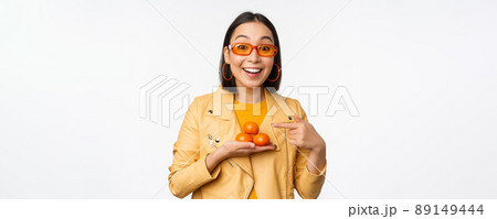 Stylish happy asian girl in sunglasses holding tangerines and smiling, posing against white background 89149444