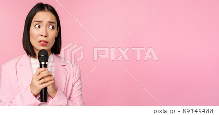 Insecure asian businesswoman giving speech, scared of talking in public, using microphone, standing in suit over pink background 89149488