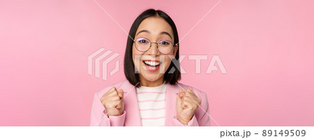 Close up portrait of asian businesswoman cheering, rooting for, looking with hope and excitement at camera, smiling and laughing, standing over pink background 89149509