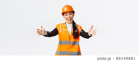 Friendly smiling asian female construction manager, engineer in safety helmet and reflective jacket, extending hands for greeting, welcome business partners, standing white background 89149881